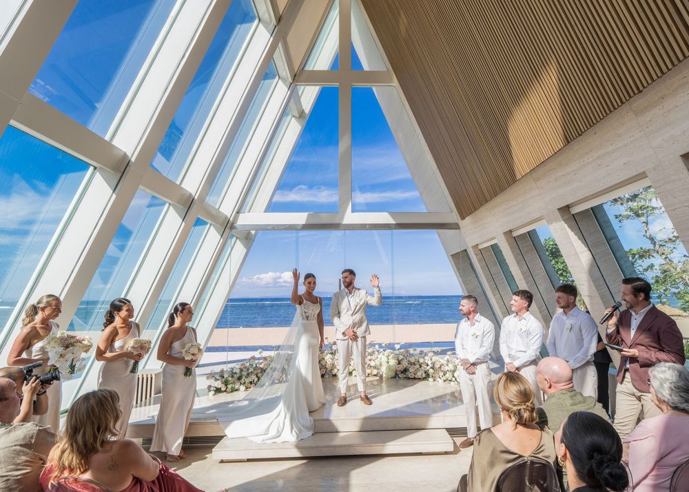 Wedding ceremony taking place in a modern chapel with soaring glass windows overlooking the ocean on a sunny day.