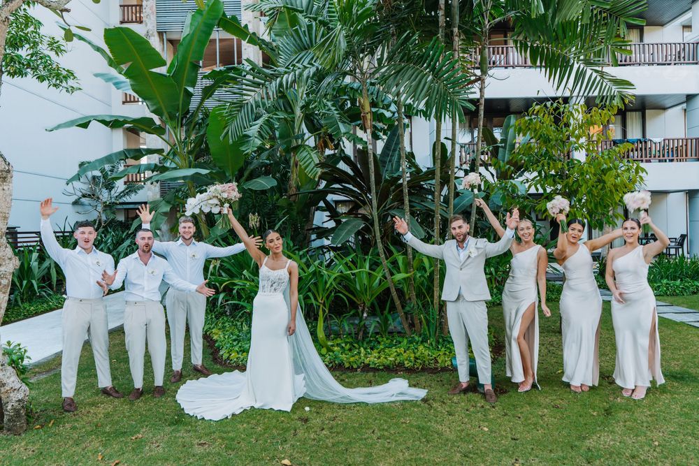 Wedding party poses under palm trees in flowing white outfits creating a series of fun and energetic group shots at Conrad Bali Resort.
