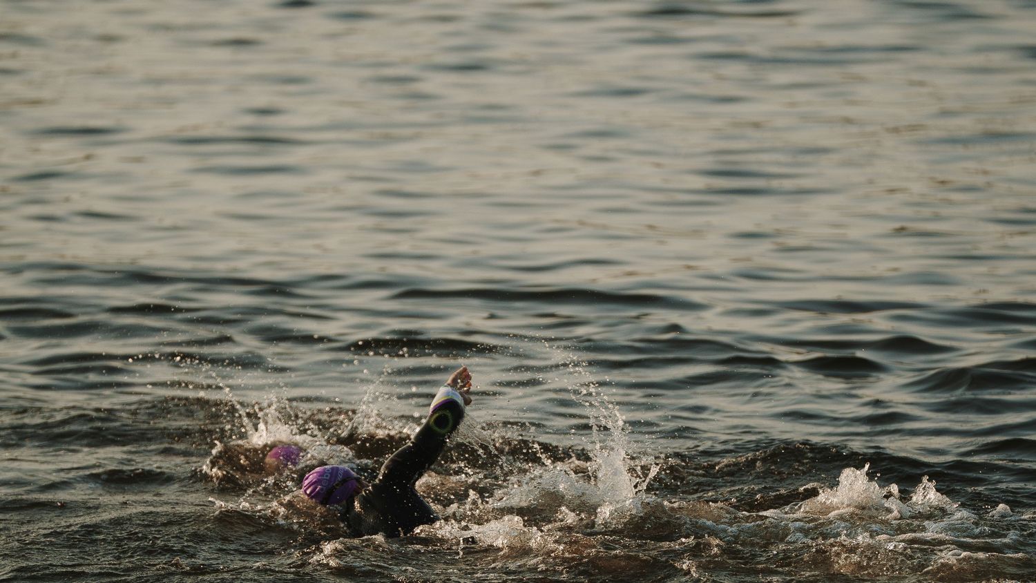A swimmer in wetsuit cutting through dark rippling water with splashing motion during an open water swim.