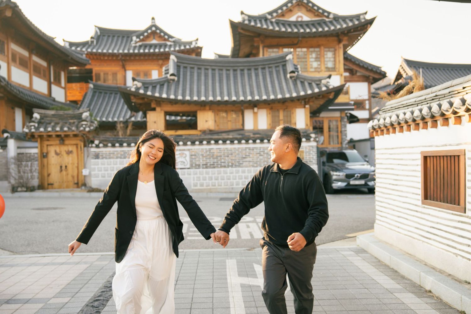 Couple strolling hand in hand through traditional Korean hanok village courtyard with ornate wooden buildings.