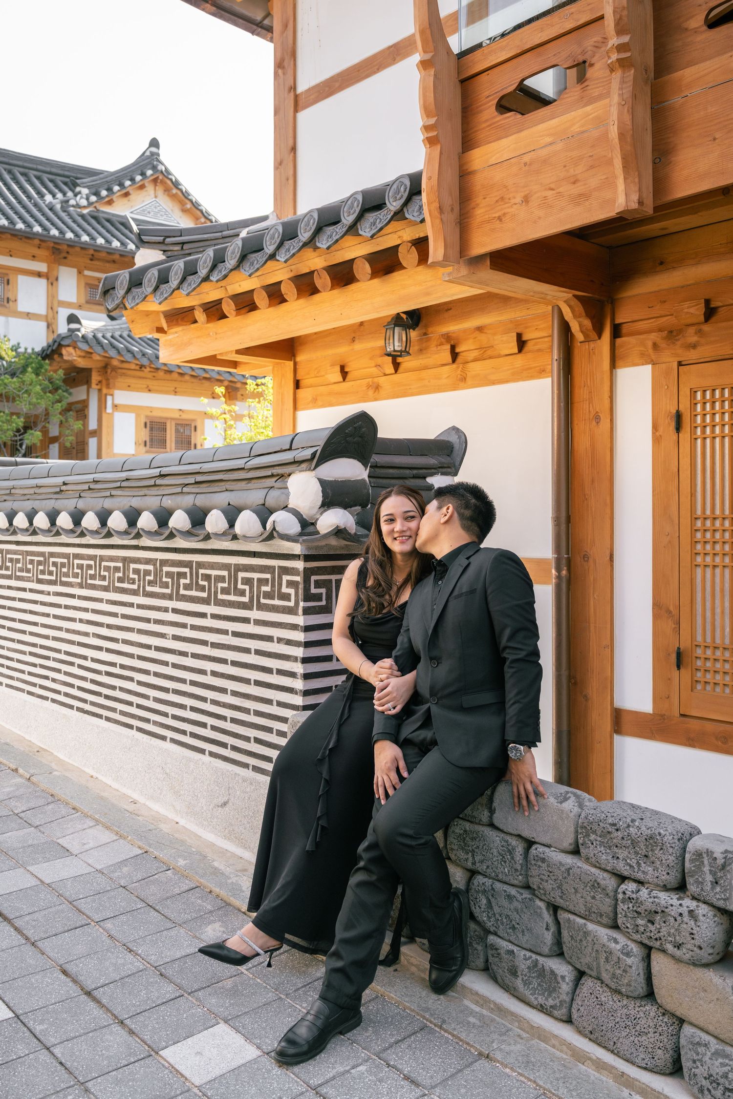 Couple in black formal attire posing together against traditional Asian architecture with stone walls and wooden beams.