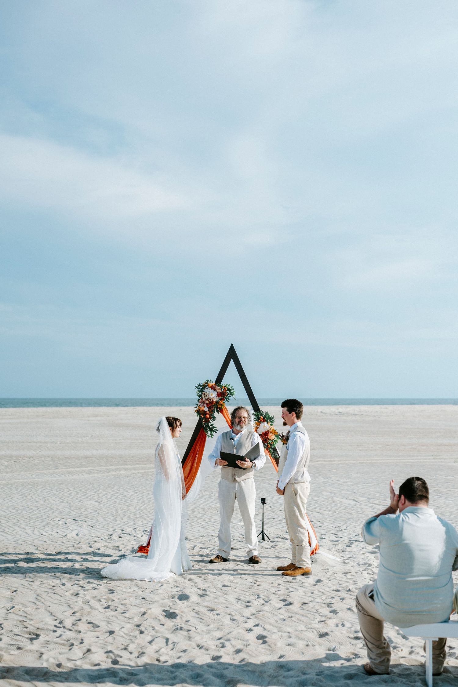 A beachside wedding ceremony takes place under a triangular arch on white sand with the ocean in the background.
