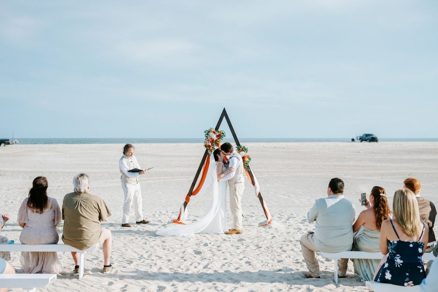 A small intimate wedding ceremony takes place on a serene white sandy beach with a wooden triangle arch and seated guests.