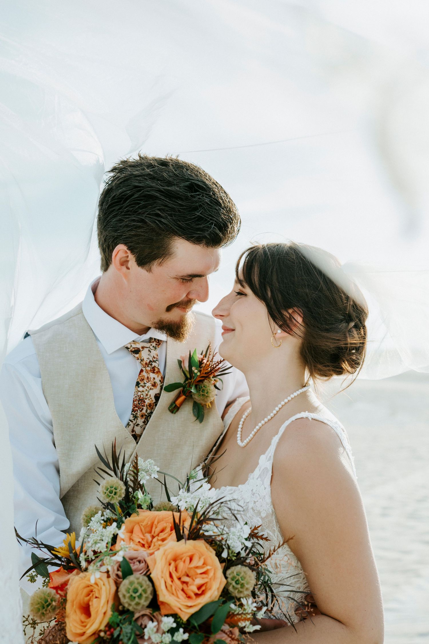 A romantic wedding couple shares an intimate moment by the beach with orange roses and white floral bouquet.