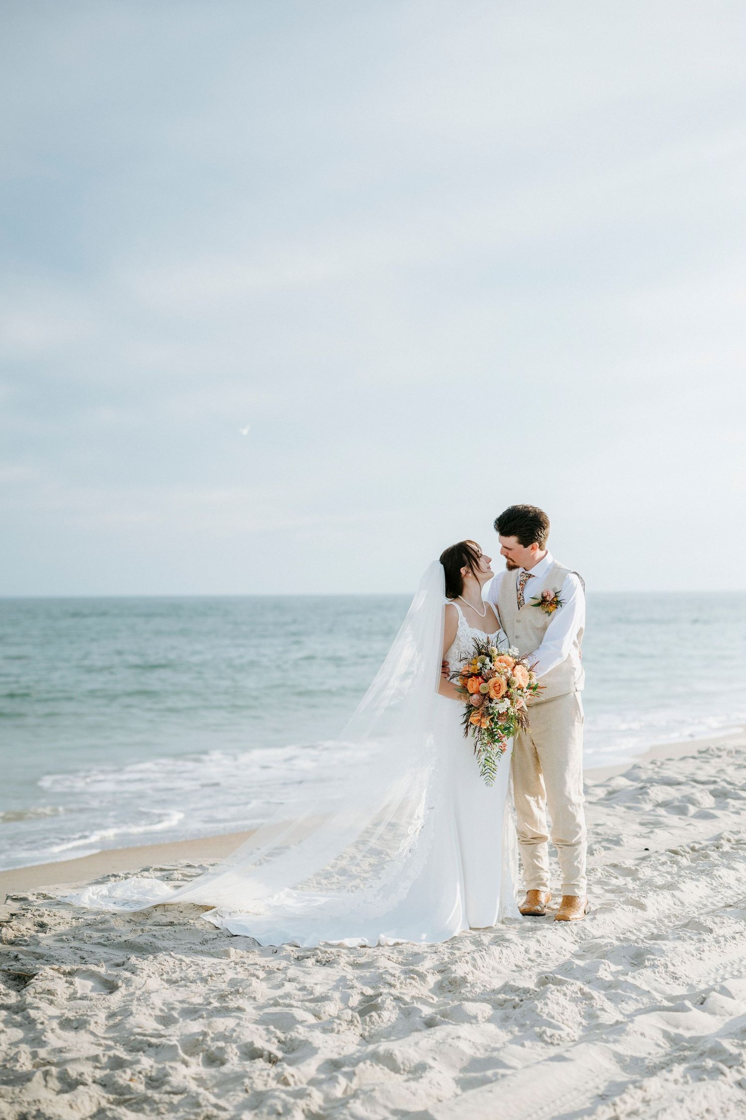 A romantic beach wedding couple embraces on white sand with flowing veil and coral bouquet against the ocean.