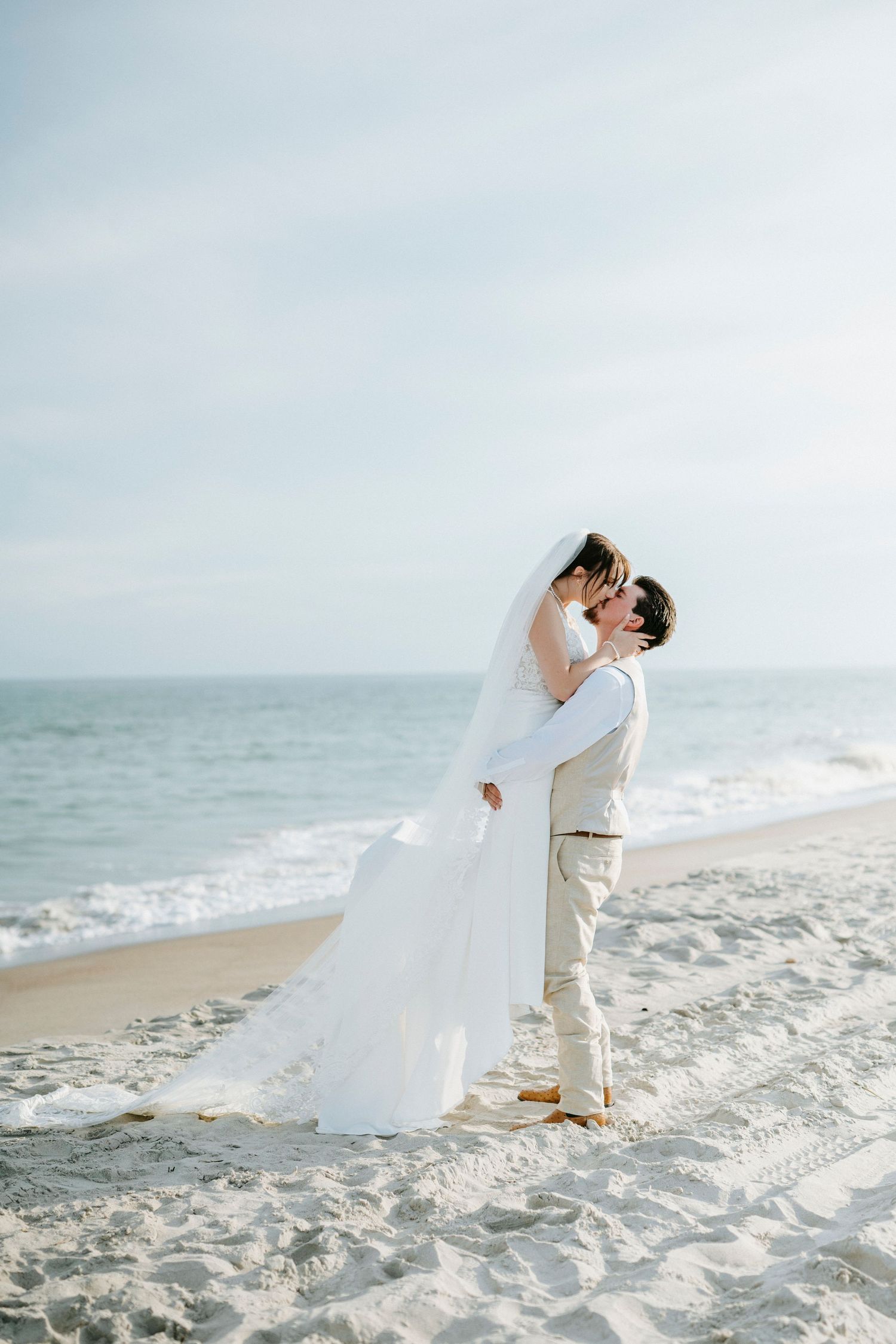 A romantic beach wedding scene with a couple embracing on white sand as waves roll in behind them.