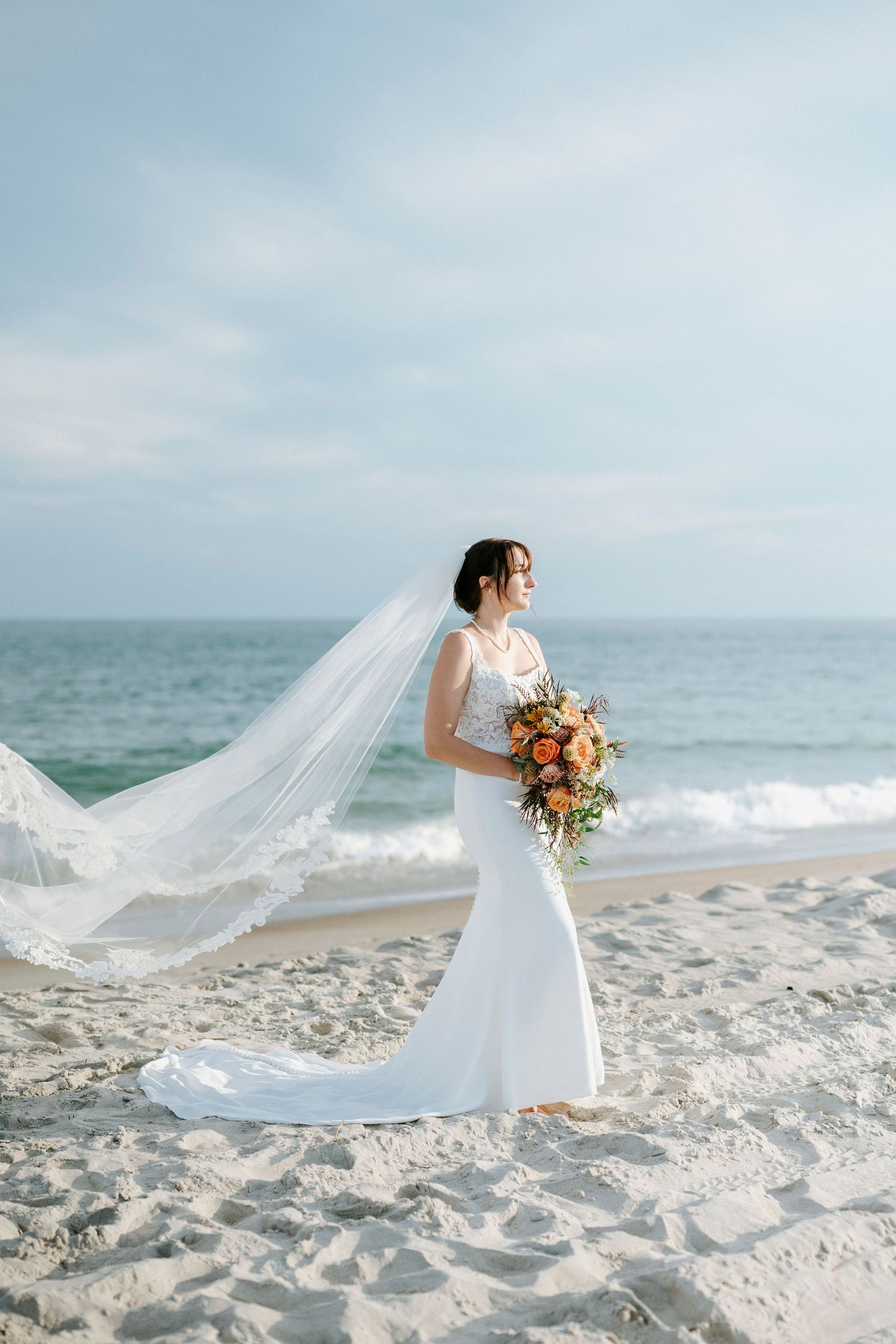 A bride in a flowing white dress walks along the beach with her veil floating in the ocean breeze.