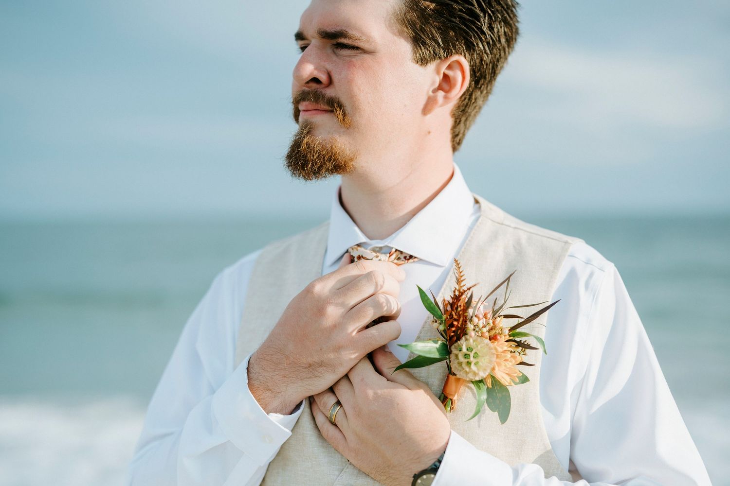 A groom in a white dress shirt adjusts his bowtie with flowers pinned to his lapel by the ocean.