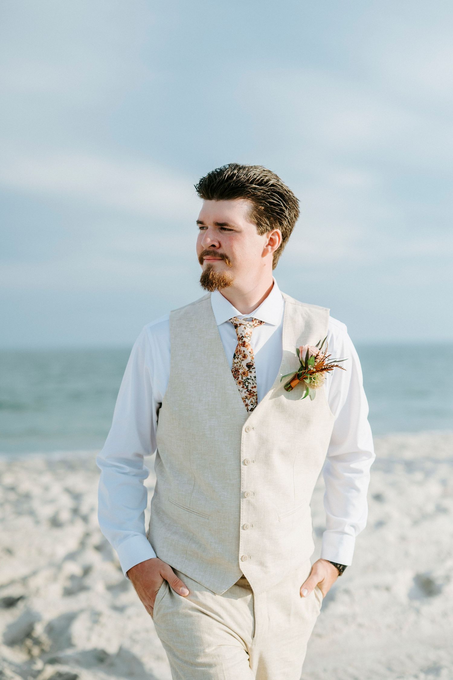 Groom in light beige vest and white shirt stands on beach with boutonniere against ocean backdrop.