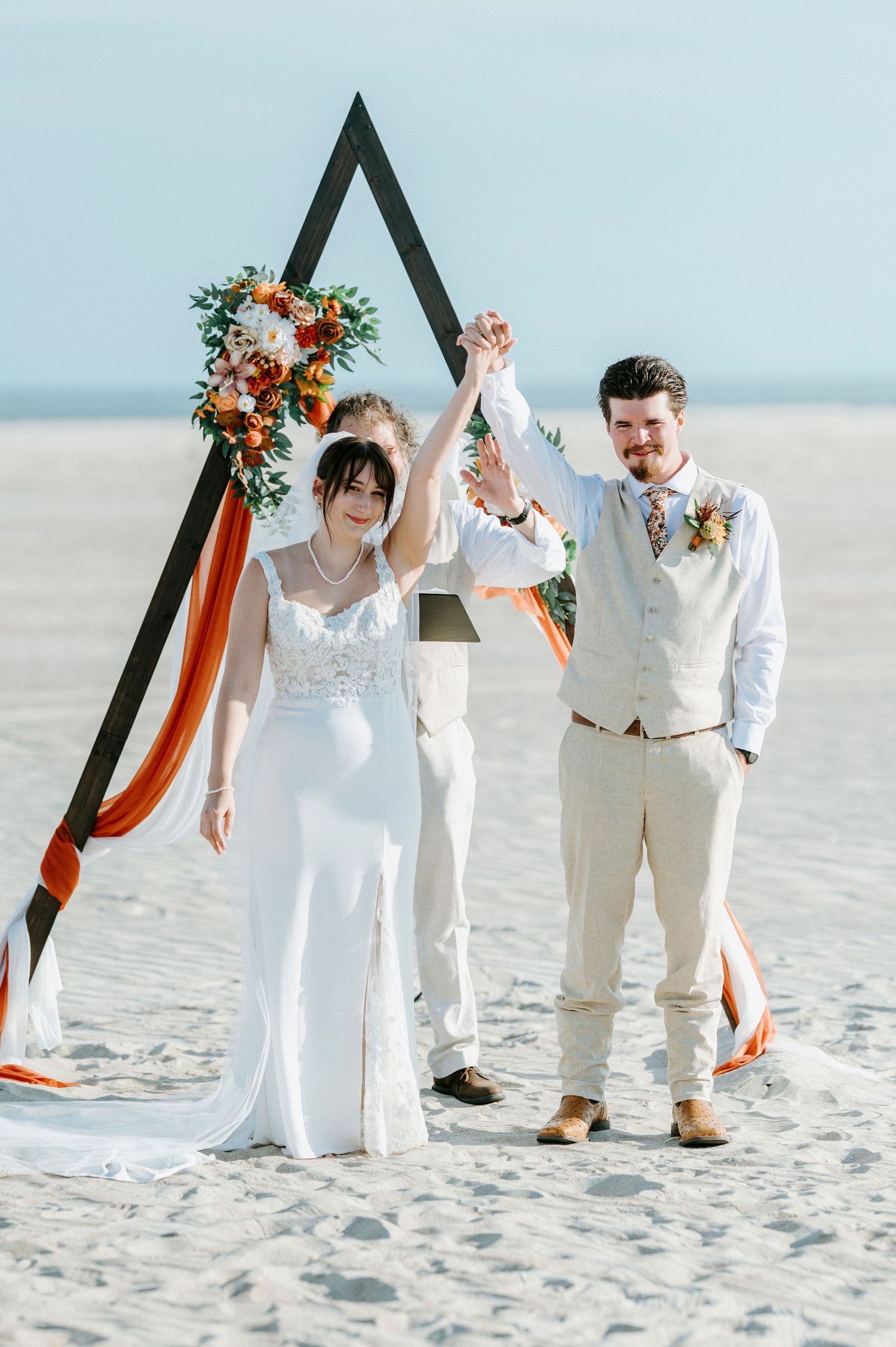 Newlyweds celebrate under triangular arch decorated with orange florals on sunny beach.