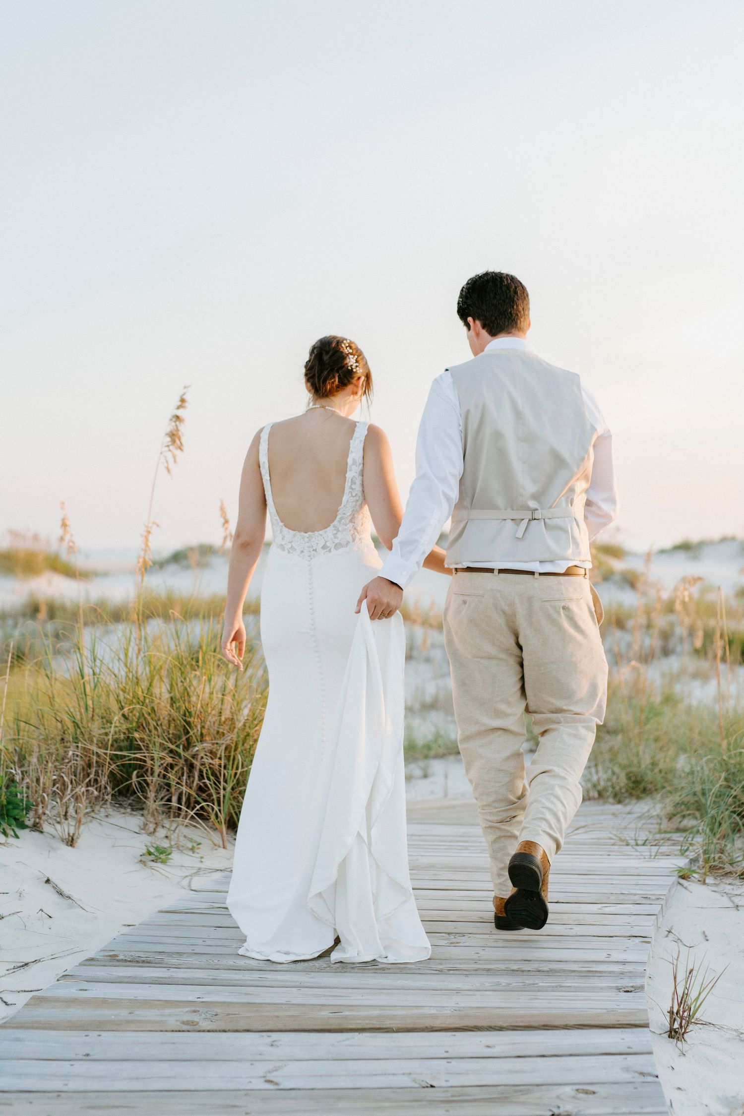 Newlyweds walk hand in hand on wooden boardwalk through beach dunes at sunset.