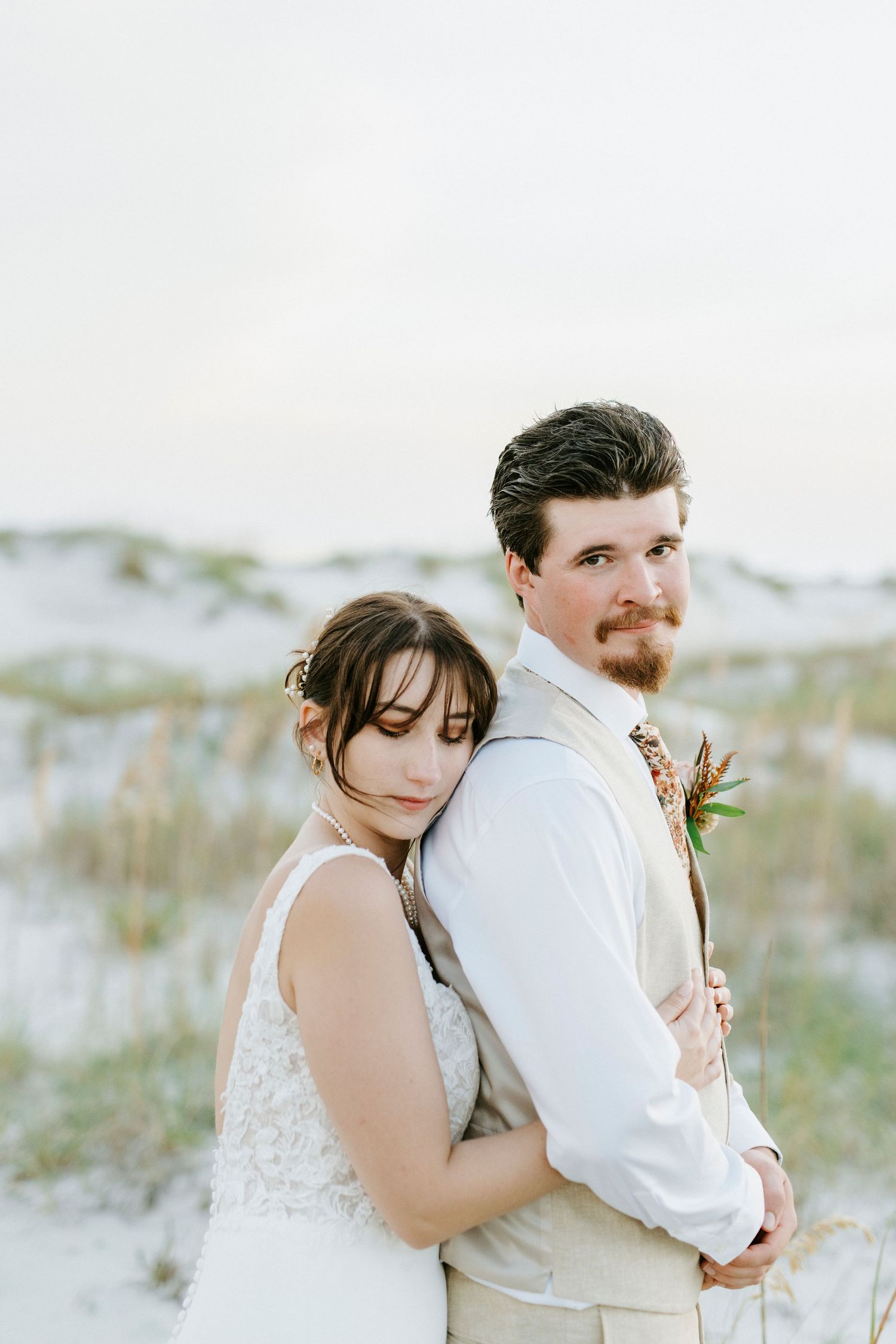 A romantic beach wedding couple embraces against the sandy dunes in soft natural lighting.