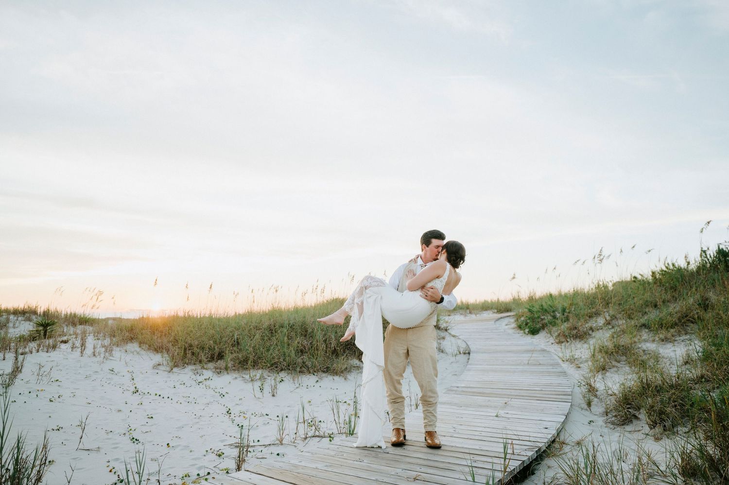 Romantic coastal photoshoot of couple walking on sandy beach boardwalk during sunset, dressed in soft neutral colors.