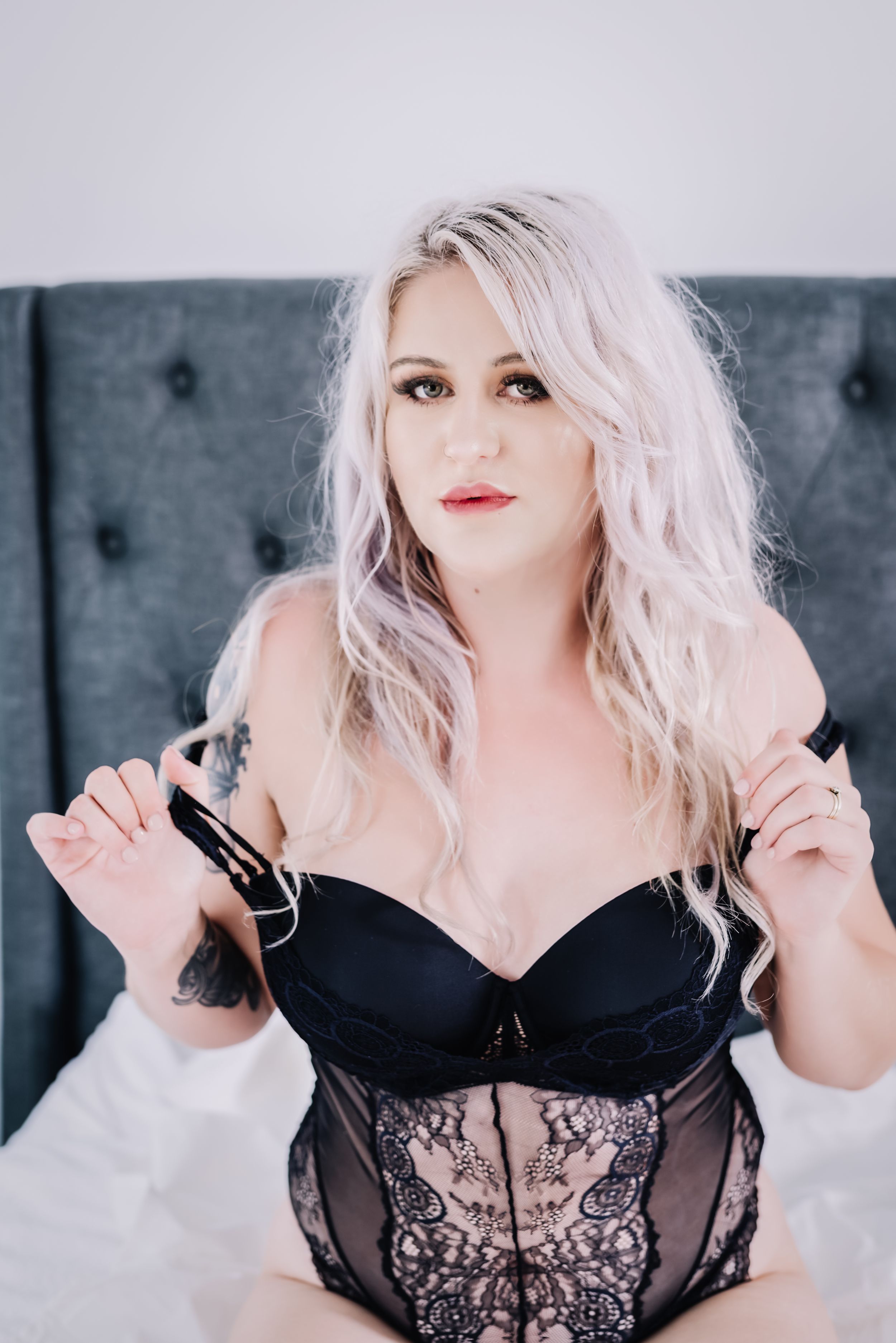 Woman with platinum blonde hair wearing black lace lingerie against grey tufted headboard.