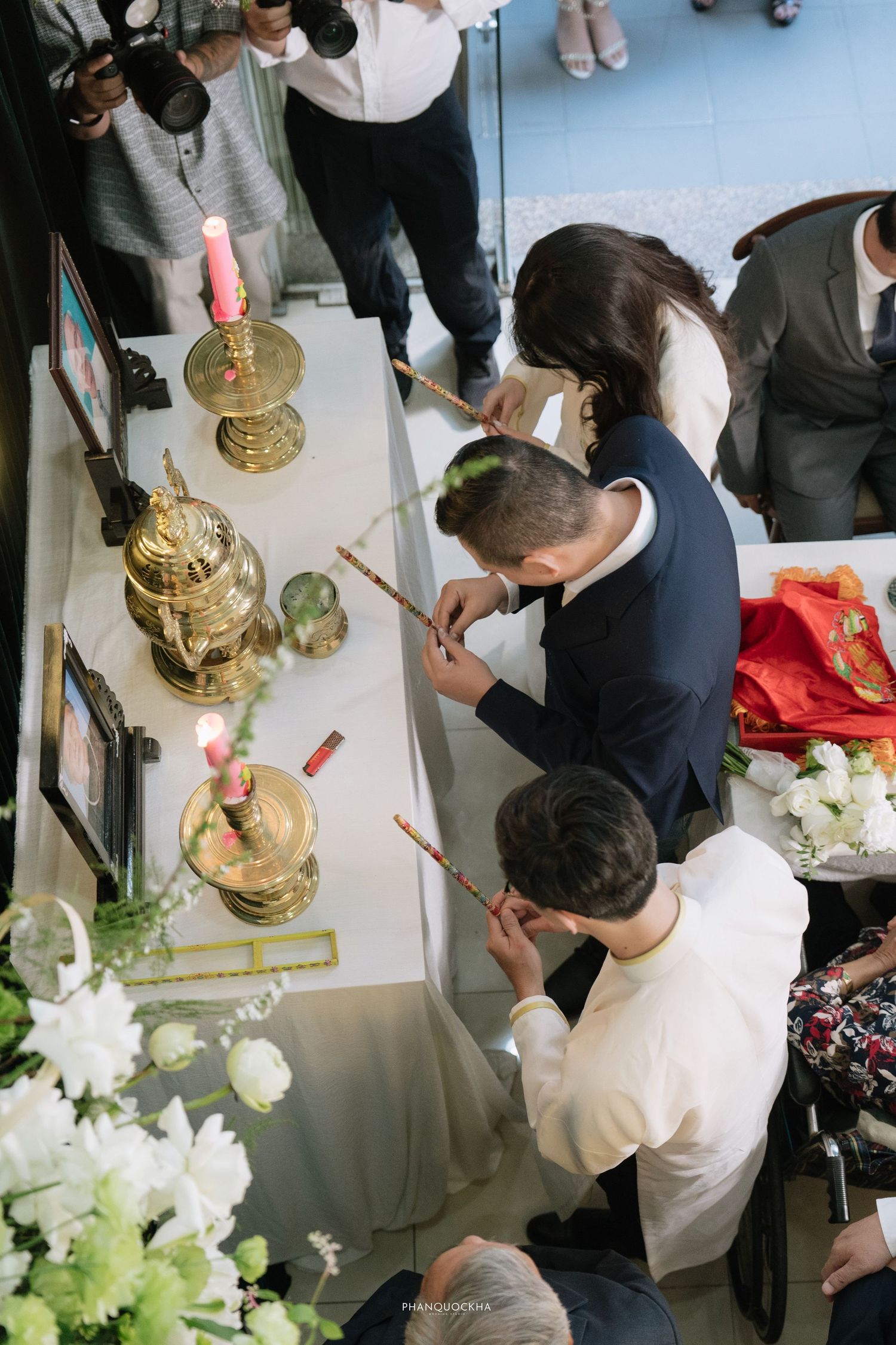 Overhead view of people offering incense at a brass ceremonial altar with pink candles and flowers.