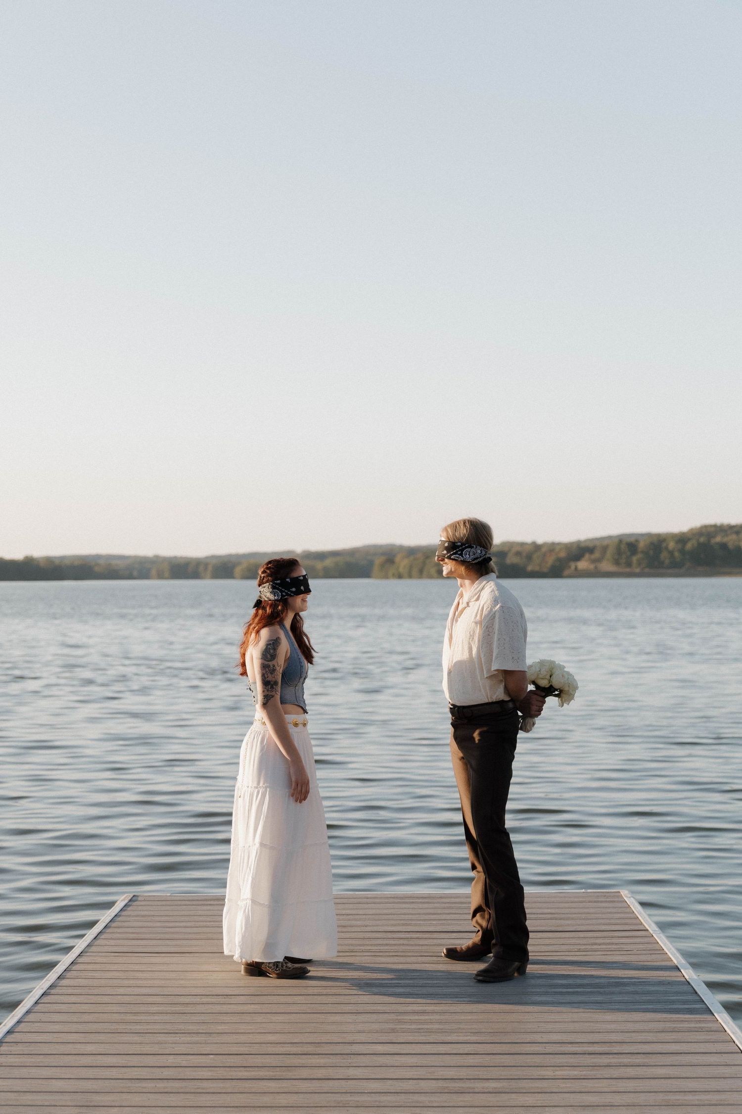 Two people in formal attire stand on a wooden dock overlooking a serene lake at sunset.