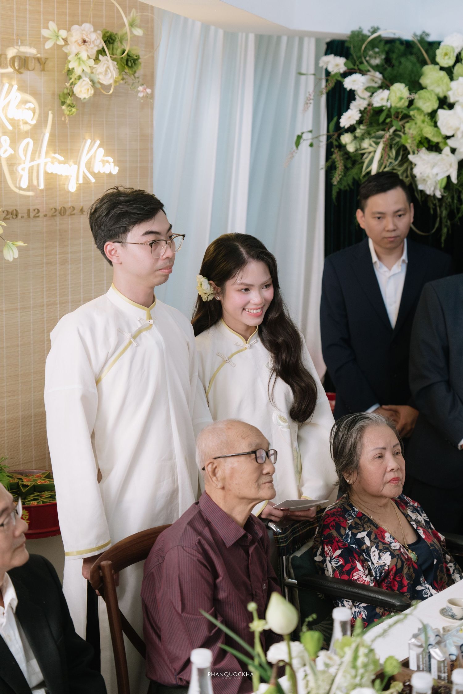 Young couple in white ao dai smiling while greeting elderly family members at a Vietnamese ceremony.