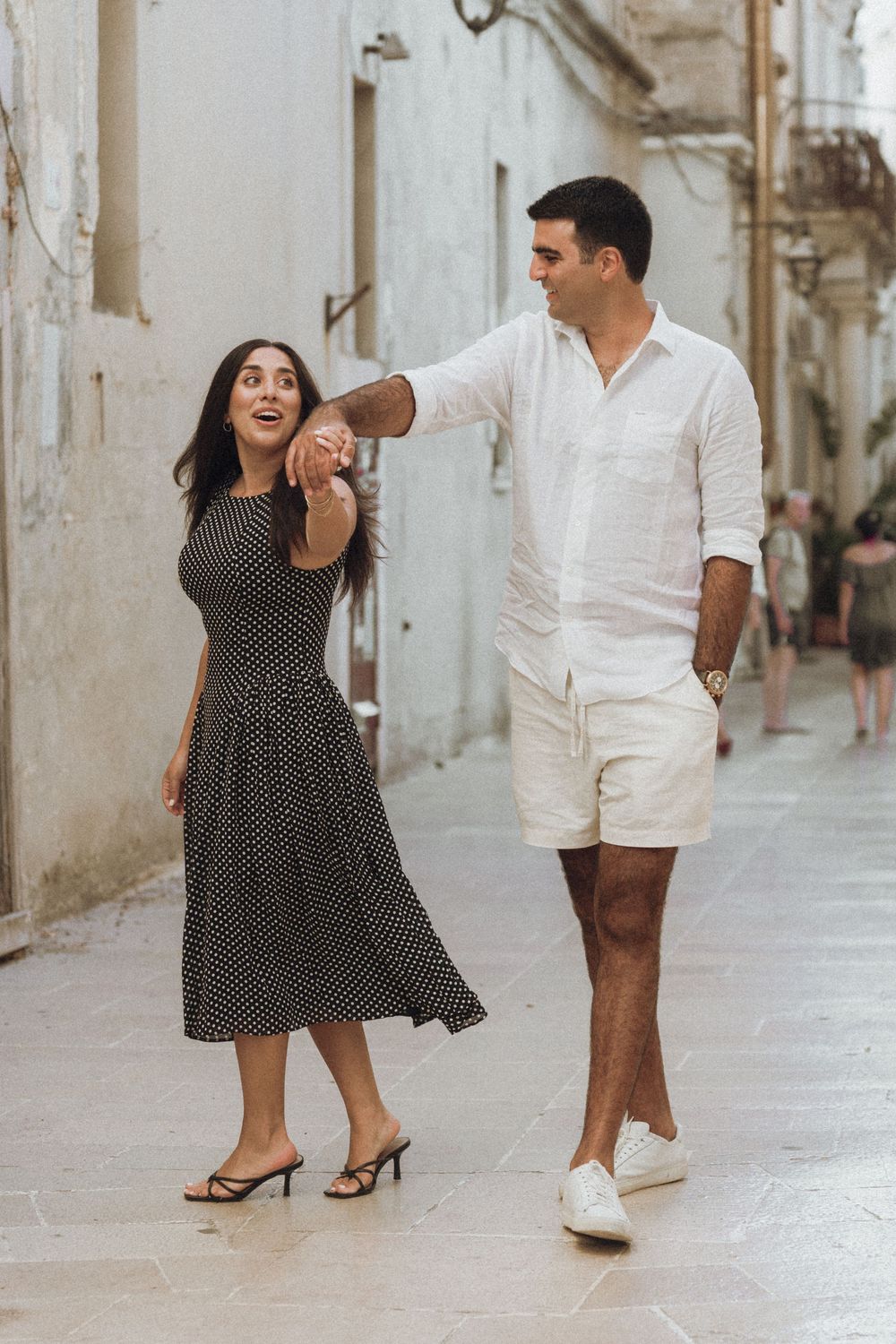 Couple in polka dot dress and white shirt dancing playfully in stone courtyard during romantic photoshoot.