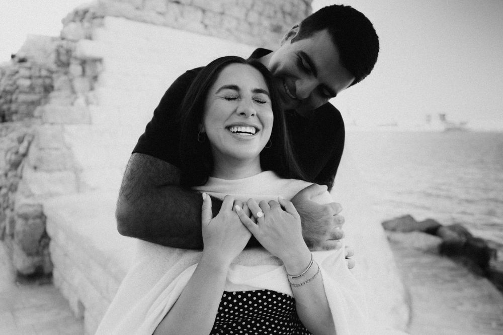 A romantic beach photoshoot in black and white showing a couple embracing by the ocean waves with cliffs nearby.