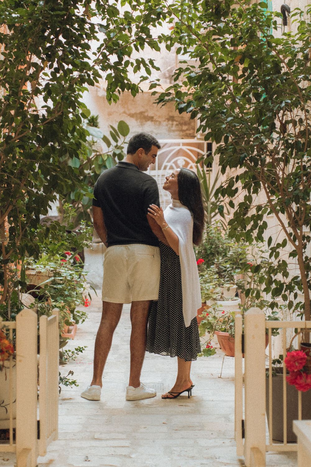A couple shares a tender moment in an outdoor garden walkway lined with trees and red flowers.