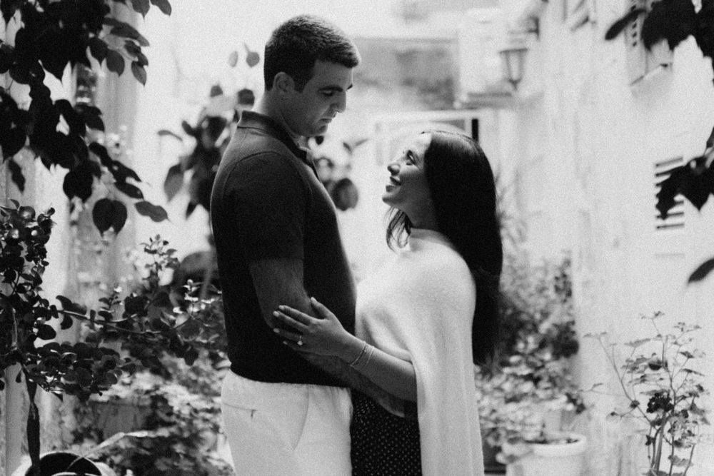 A romantic black and white photograph of a couple standing close together in a garden setting with leaves framing the scene.