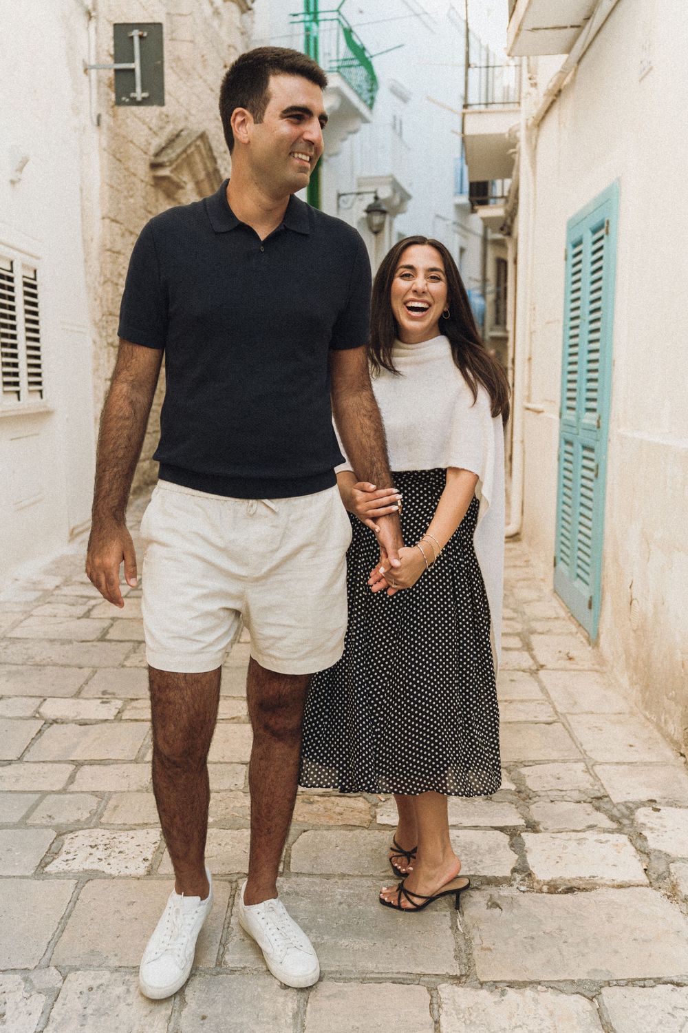 A couple walks and laughs together along charming Mediterranean alleyways with white stone walls and turquoise shutters.