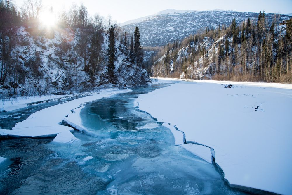 Snow-covered winter landscape with evergreen trees surrounding a winding turquoise river flowing through mountains.