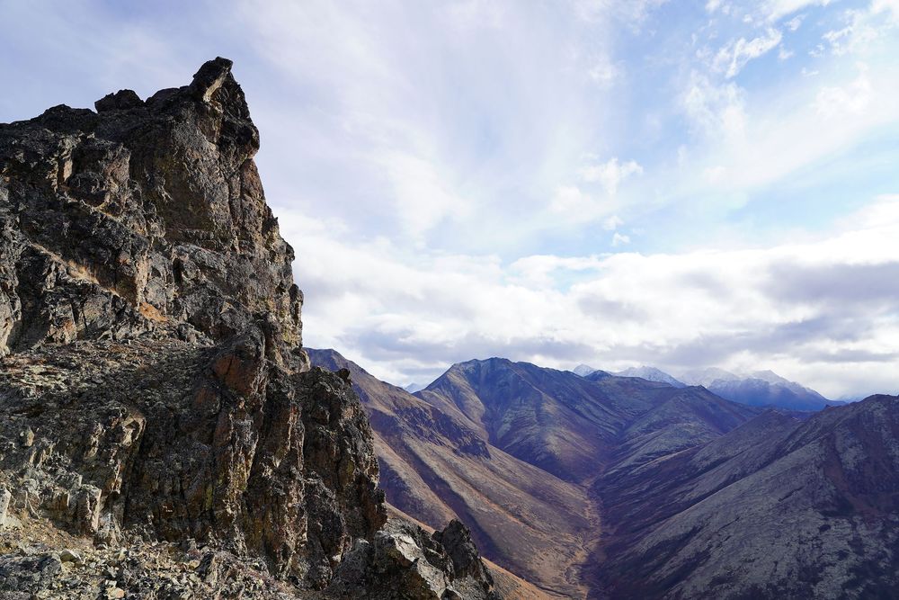 Rugged rocky cliff overlooks a deep mountain valley with distant peaks under cloudy skies in remote wilderness terrain.