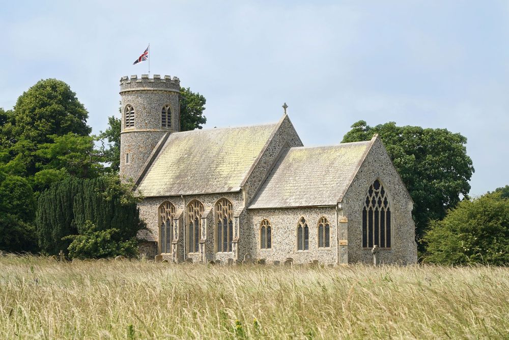 Historic stone church with round tower and flag stands among tall grass in the English countryside.
