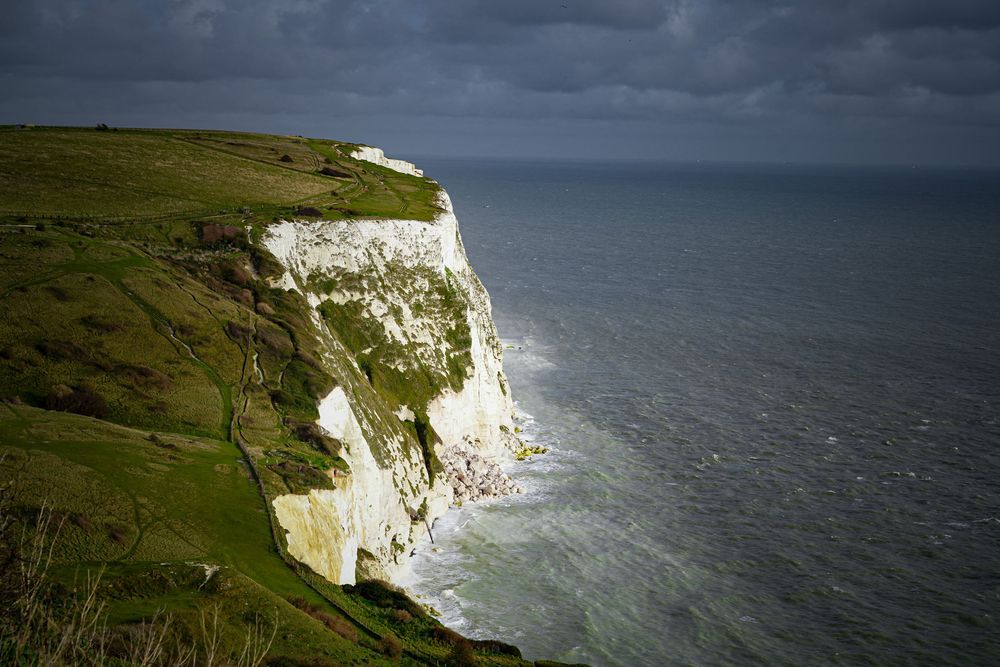 Dramatic white chalk cliffs of Dover rise above the English Channel under stormy skies with green grass on top.
