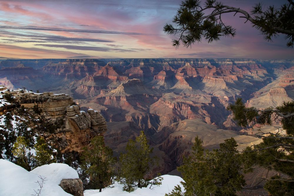 Colorful sunset illuminates snow-covered Grand Canyon vista with dramatic rock formations and evergreen trees along rim.