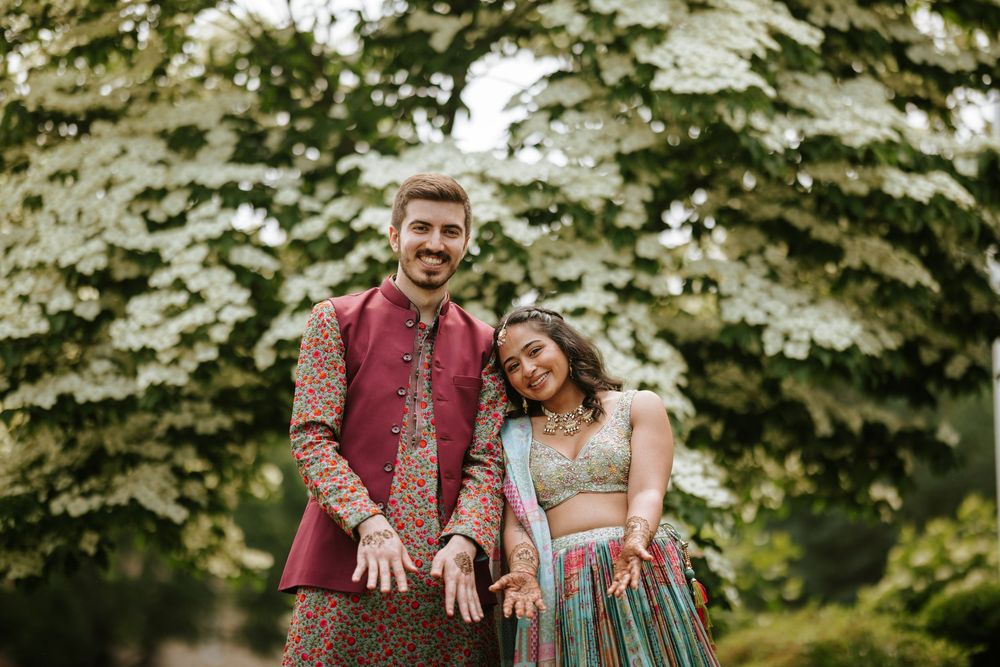 A couple in burgundy and blue outfits share joyful moments during an outdoor engagement photoshoot among evergreen trees.