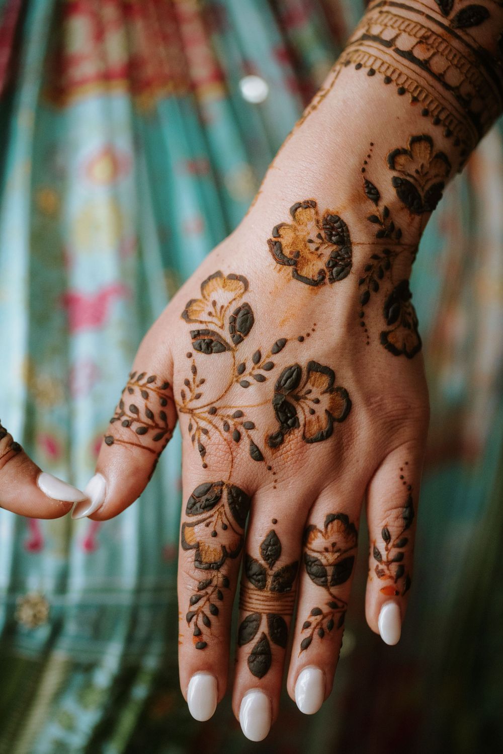 Intricate floral henna design adorns a hand with white manicured nails against a turquoise patterned fabric background.