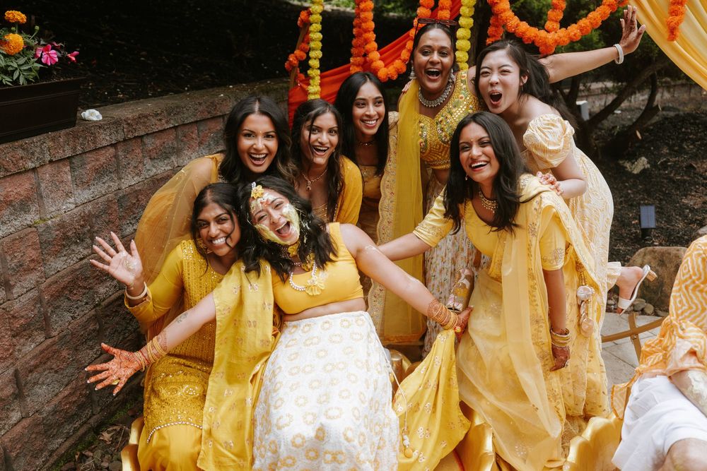 Group of friends in yellow outfits celebrating at an outdoor Indian wedding with colorful marigold decorations.