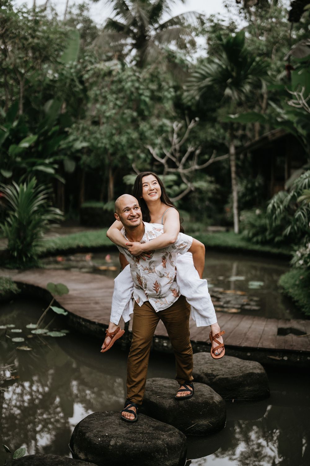 Couple posing playfully on stone steps near a reflecting pool in a tropical garden.