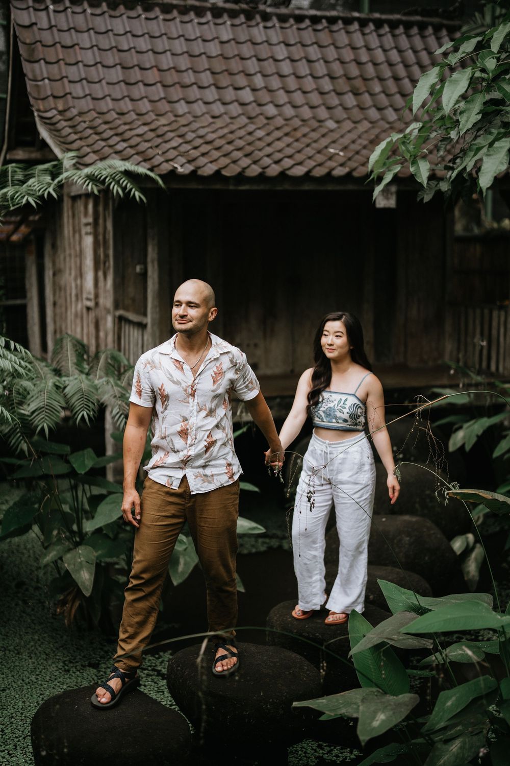 Casual couple photoshoot in front of traditional wooden buildings surrounded by tropical plants.