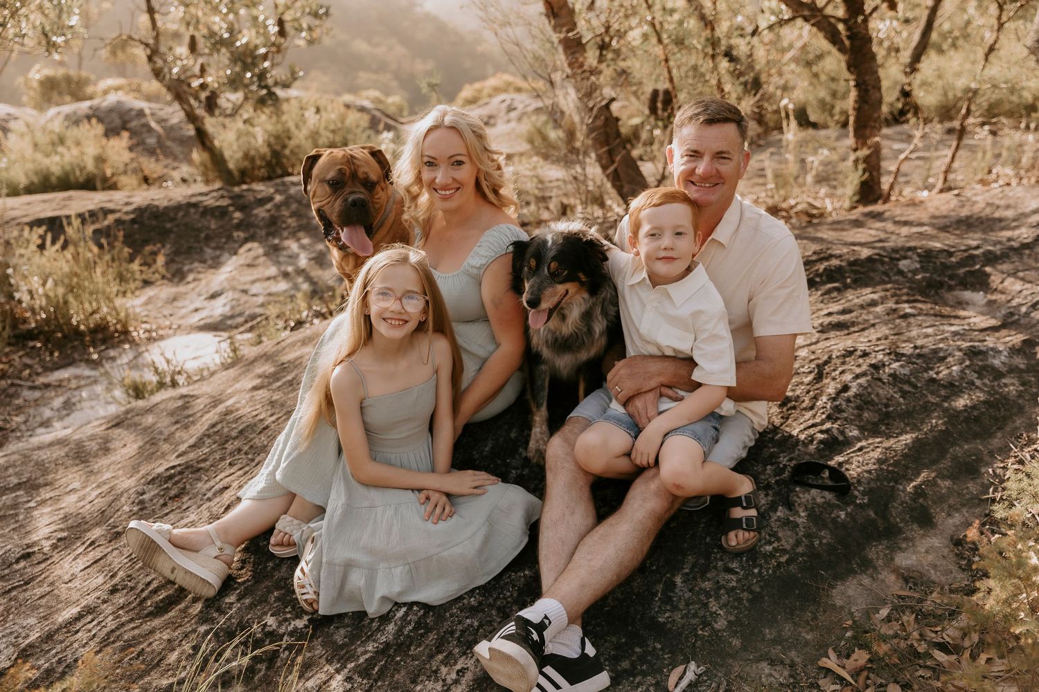 Family with children and dog sitting together outdoors in a natural wooded setting during golden hour.