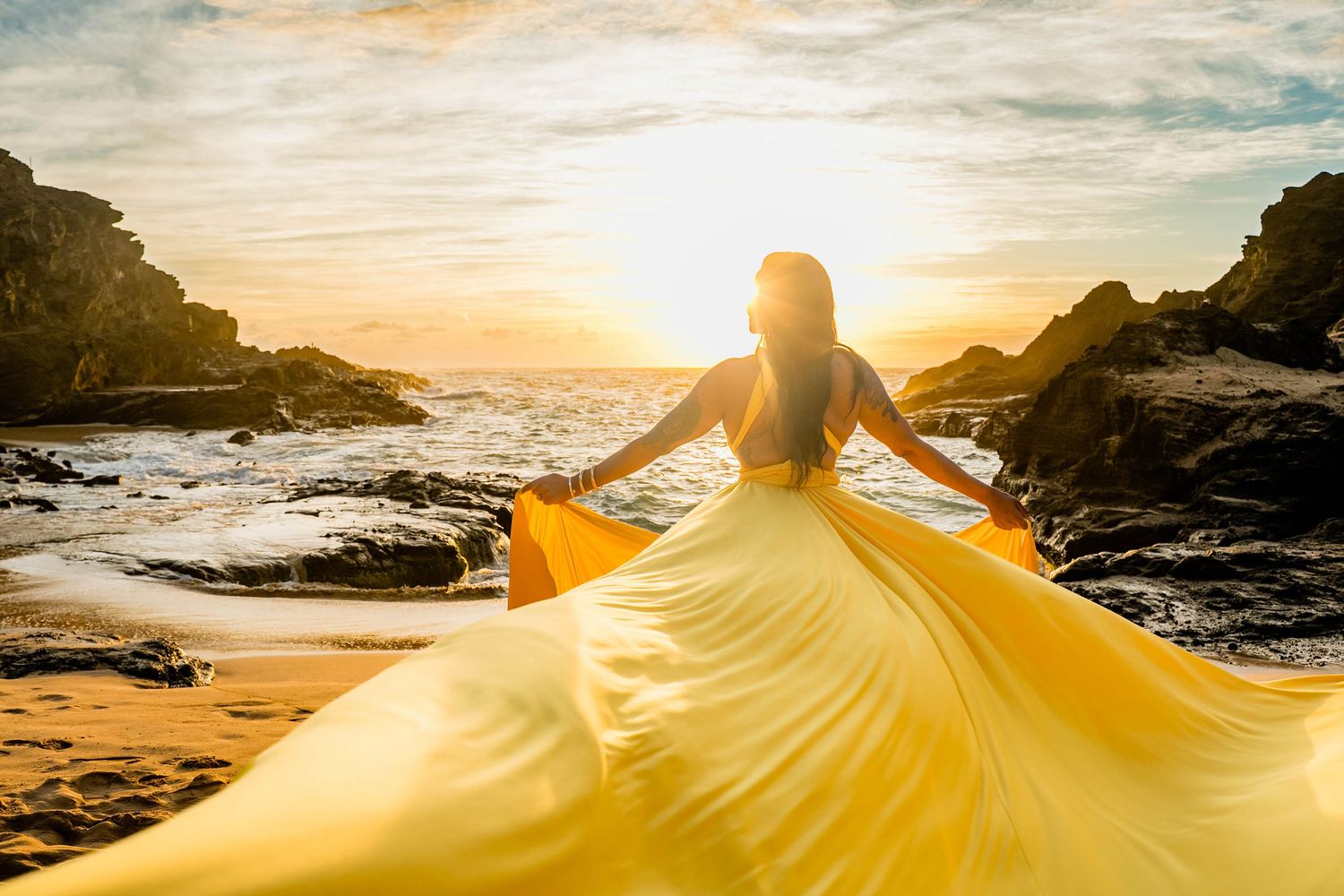 A flowing yellow dress creates a dramatic silhouette against a rocky coastal sunset landscape.