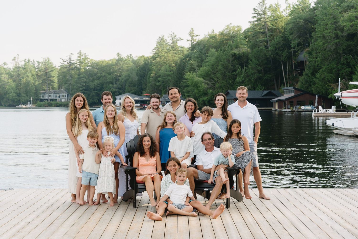 A group of people in white and beige outfits pose together on a wooden dock by a lake during summer.
