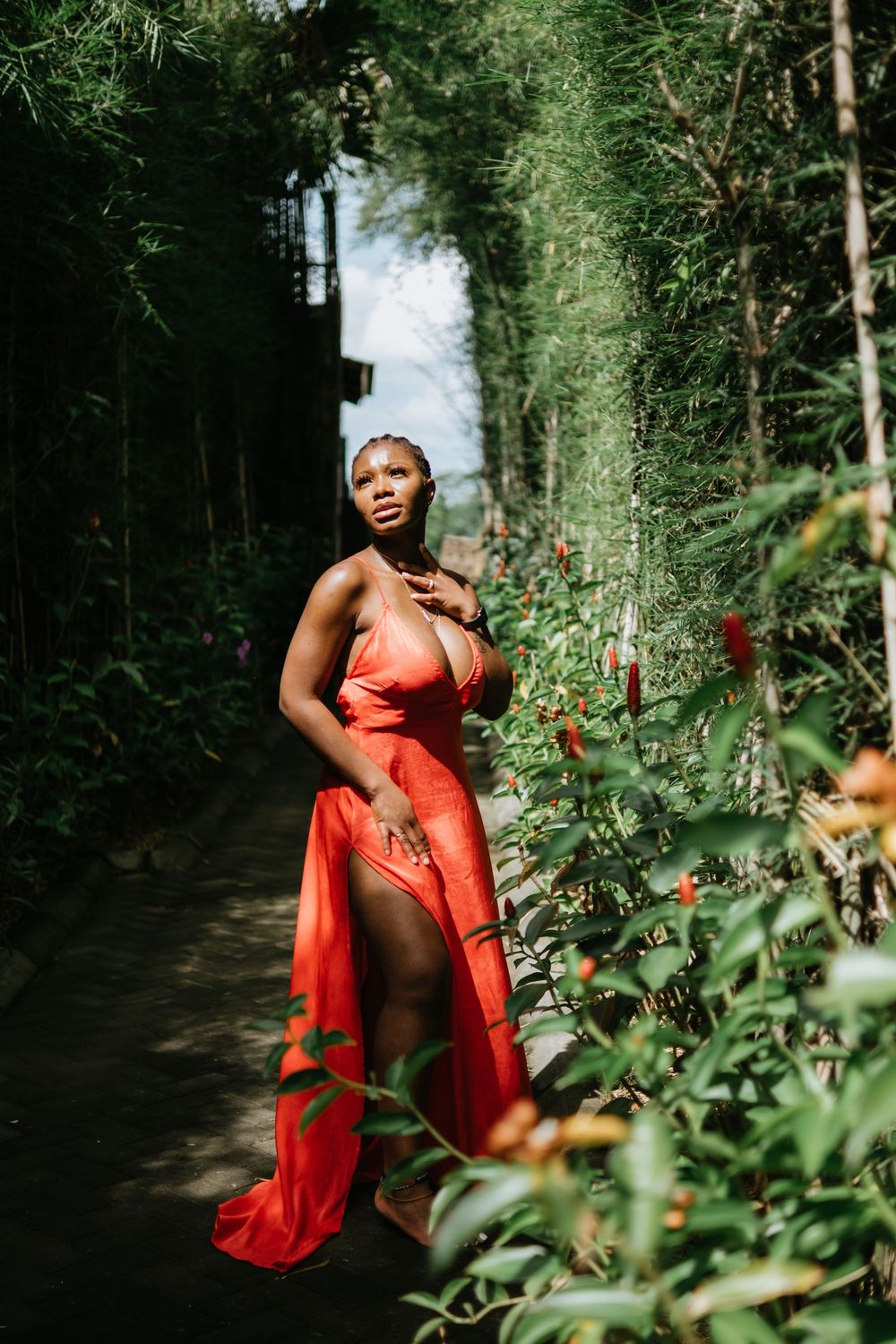 Woman in flowing red dress poses elegantly in bamboo forest pathway.