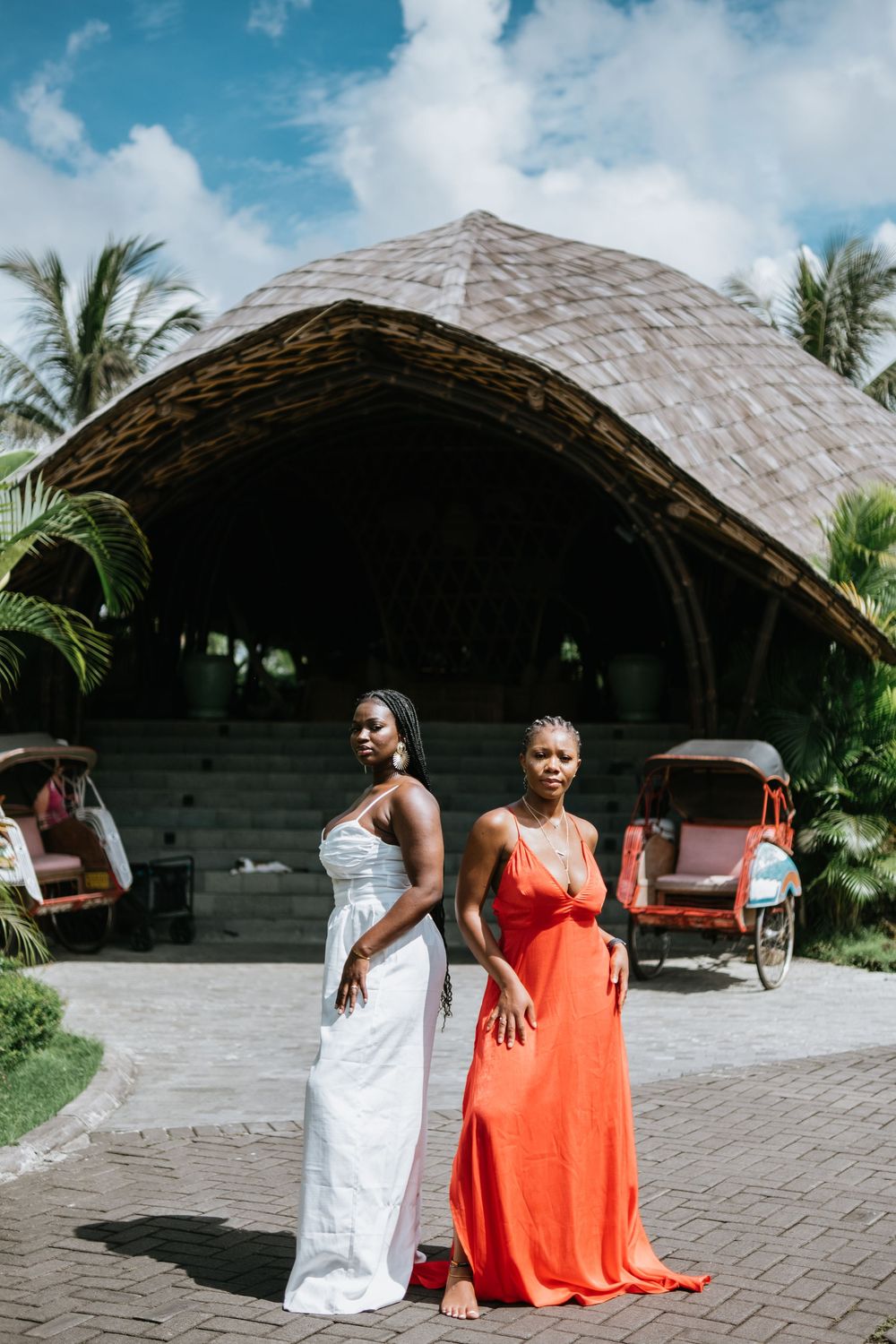 Two women pose in elegant dresses in front of a thatched-roof pavilion with palm trees in a tropical setting.