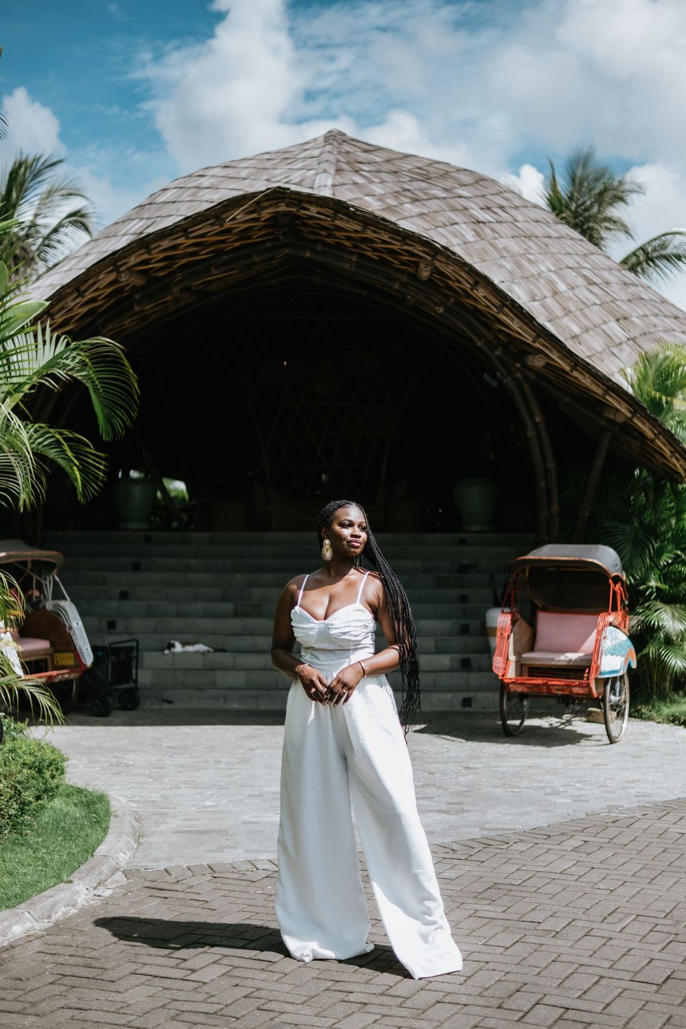 Woman in white jumpsuit poses elegantly in front of tropical pavilion with palm trees and brick path.