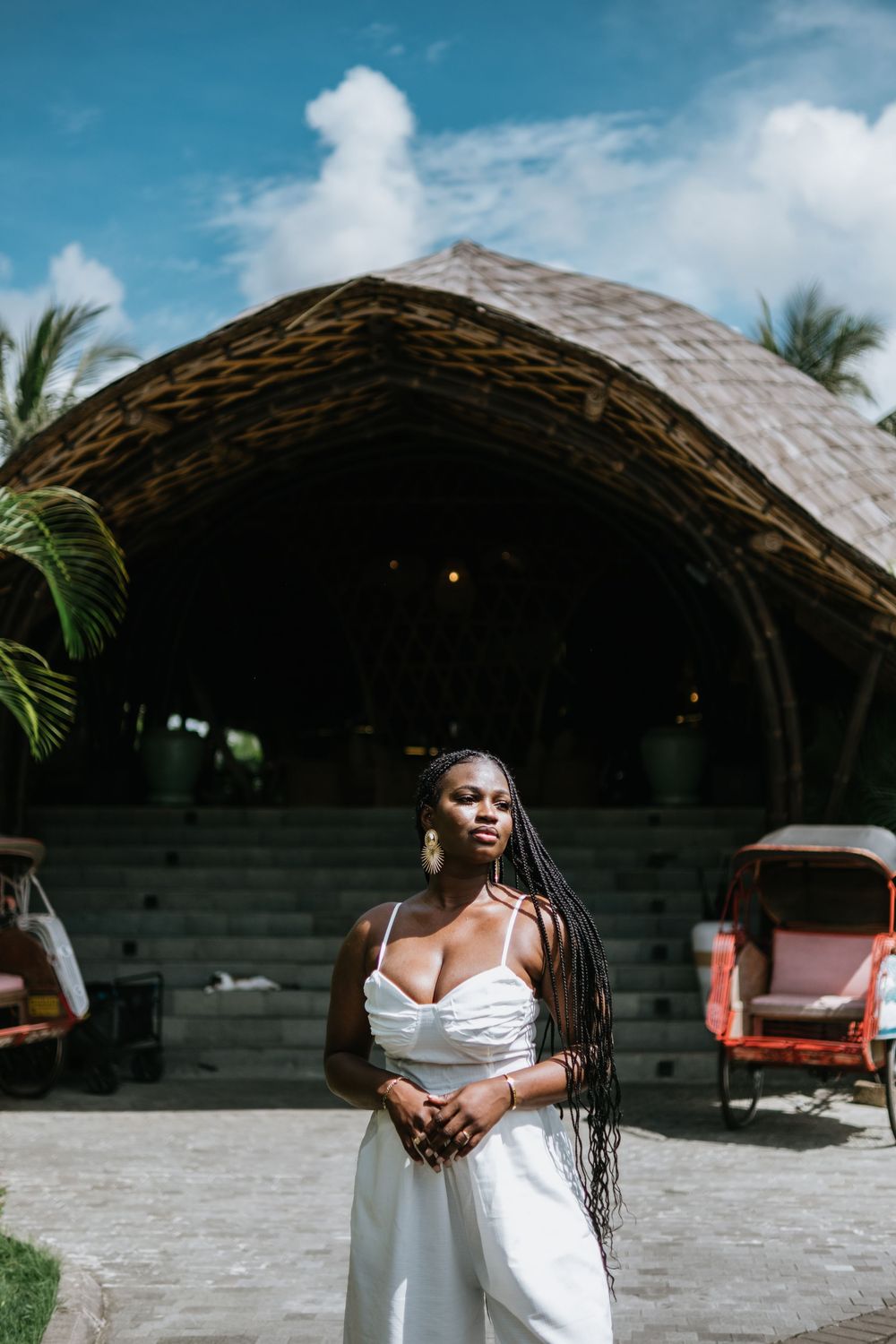 Woman in white outfit stands confidently in front of curved bamboo architecture and tropical landscaping.