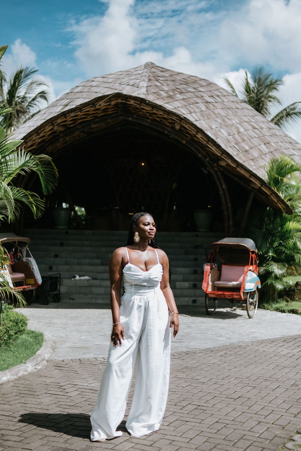 Stylish woman in white jumpsuit poses against backdrop of tropical resort structure.