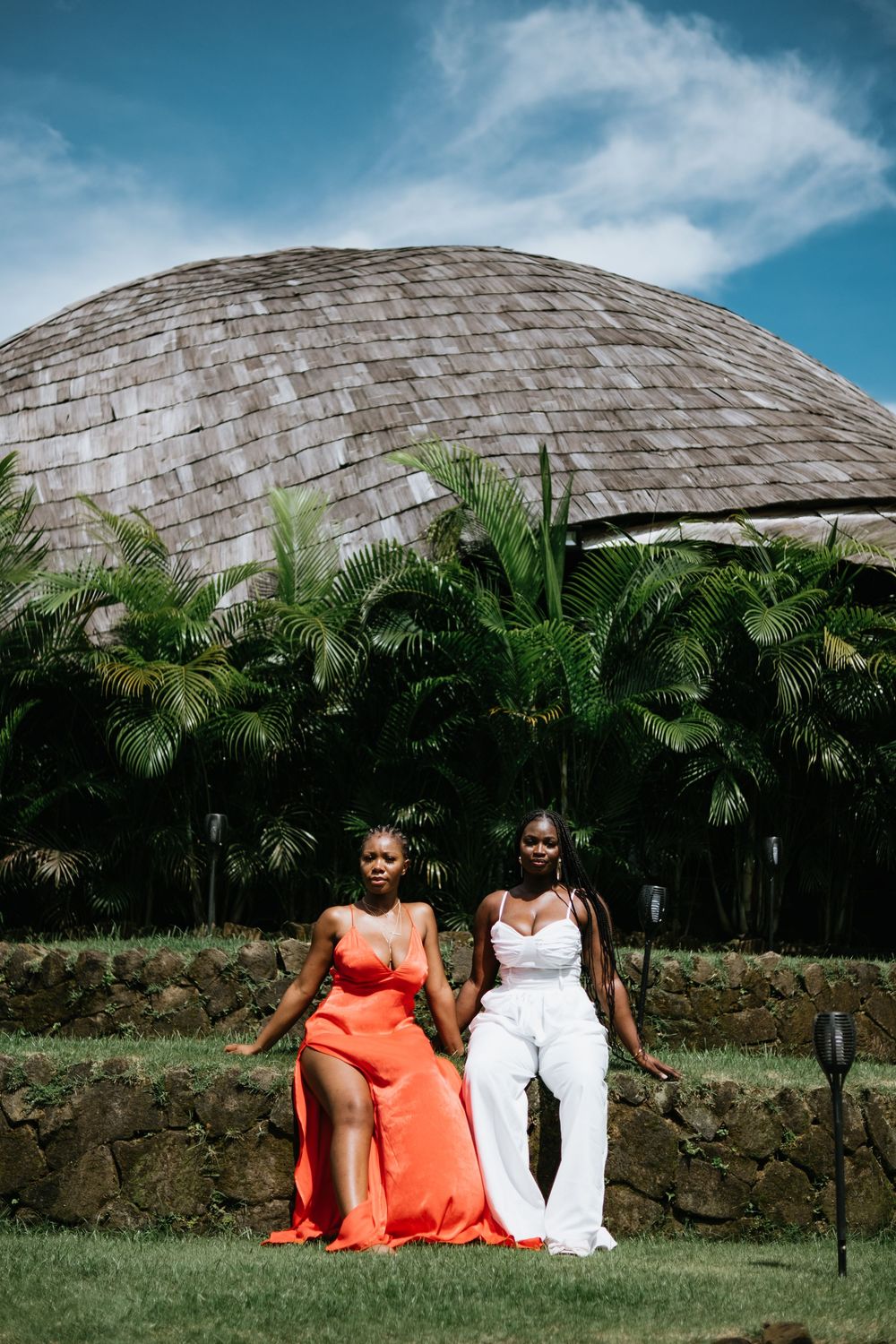Two women in orange and white outfits sit on stone wall with domed building and palms in background.