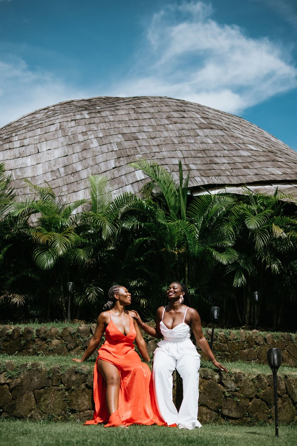 Two friends share a joyful moment on stone wall with tropical dome architecture behind them.