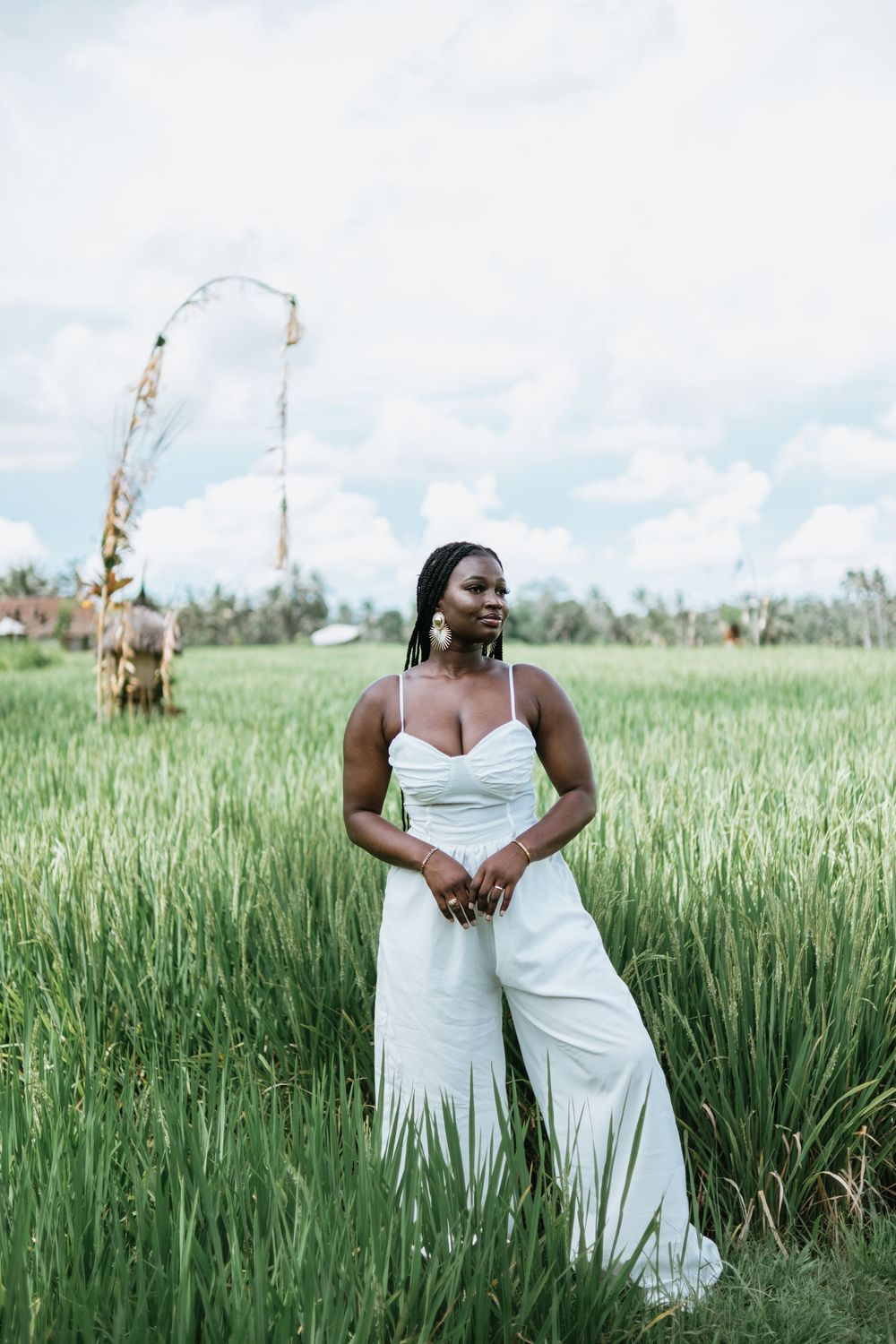 Woman in white jumpsuit poses gracefully in tall grass field with palm trees in background.