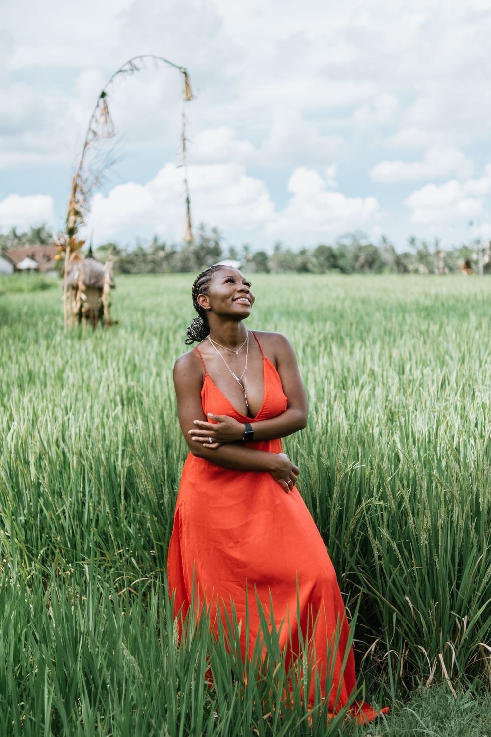 Woman in flowing coral dress stands amid lush green rice field under cloudy sky.