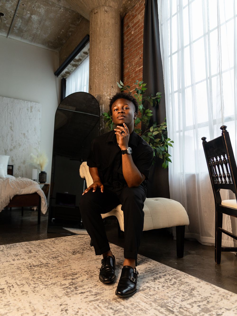 A person in black clothing sits on a wooden chair in an industrial-style room with large windows.