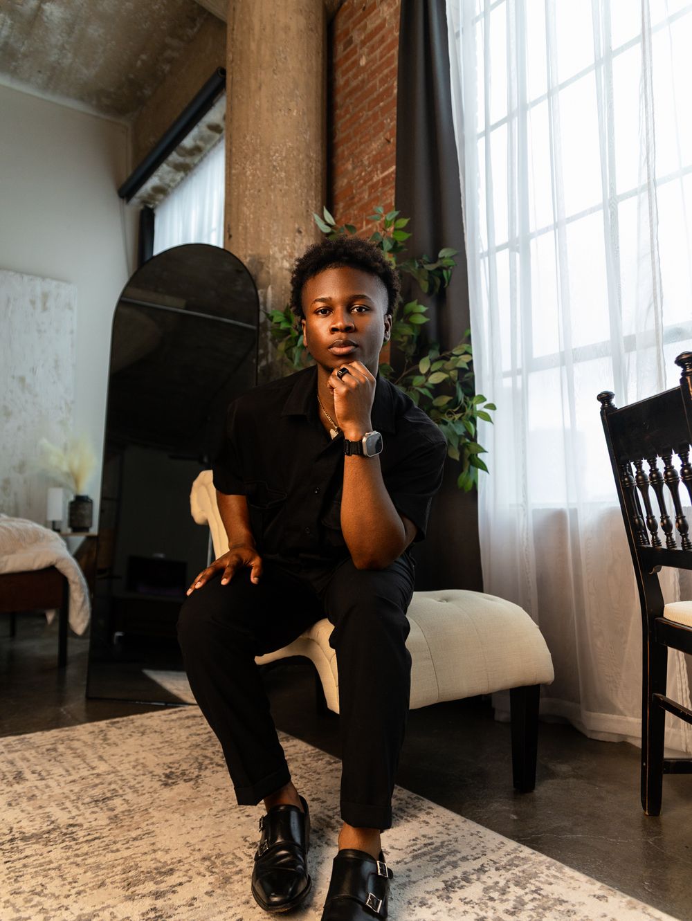 A figure in dark clothing poses on a patterned rug in a room with exposed brick walls and natural light.